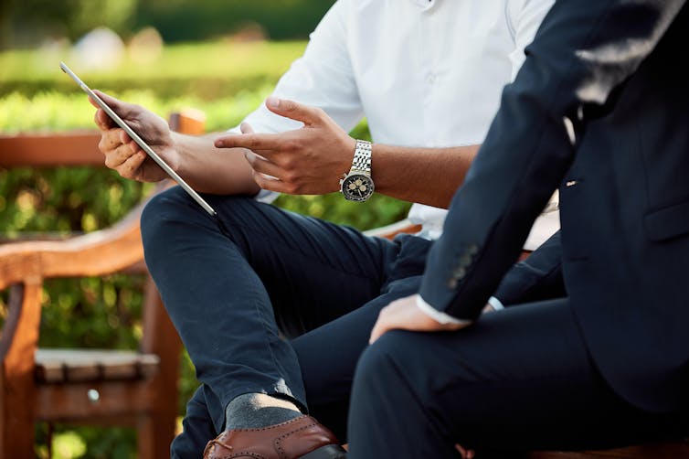A man looks at a tablet as another man sits behind him in an outdoor setting.