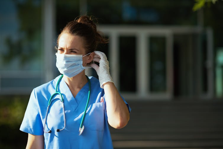 A health-care worker stands outdoors, adjusting her mask.