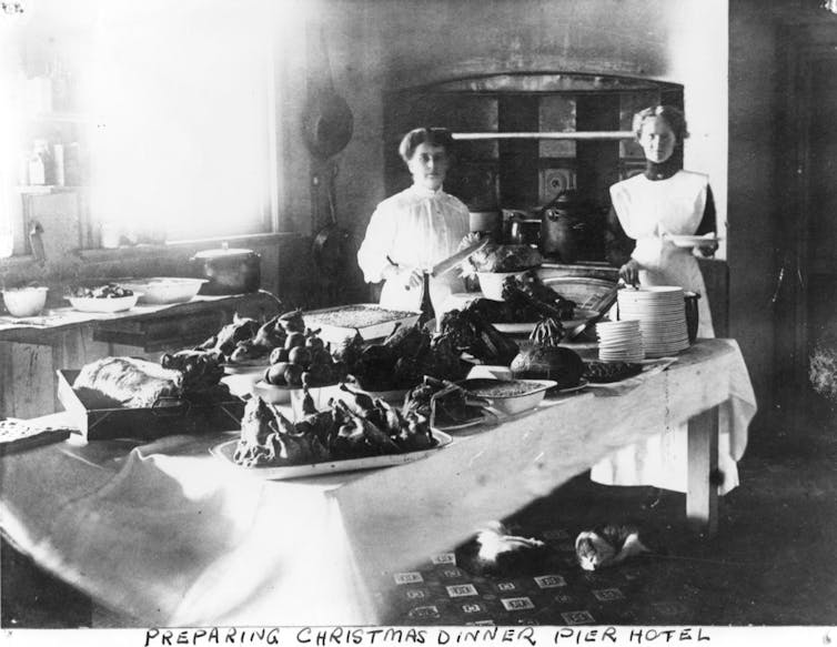 Two maids stand by a table laden with meat.