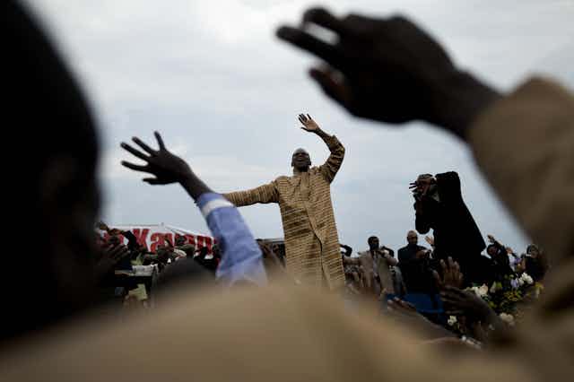 In the foreground, hands are raised in the air and beyond them, a man in traditional attire raises his own hands heavenward.