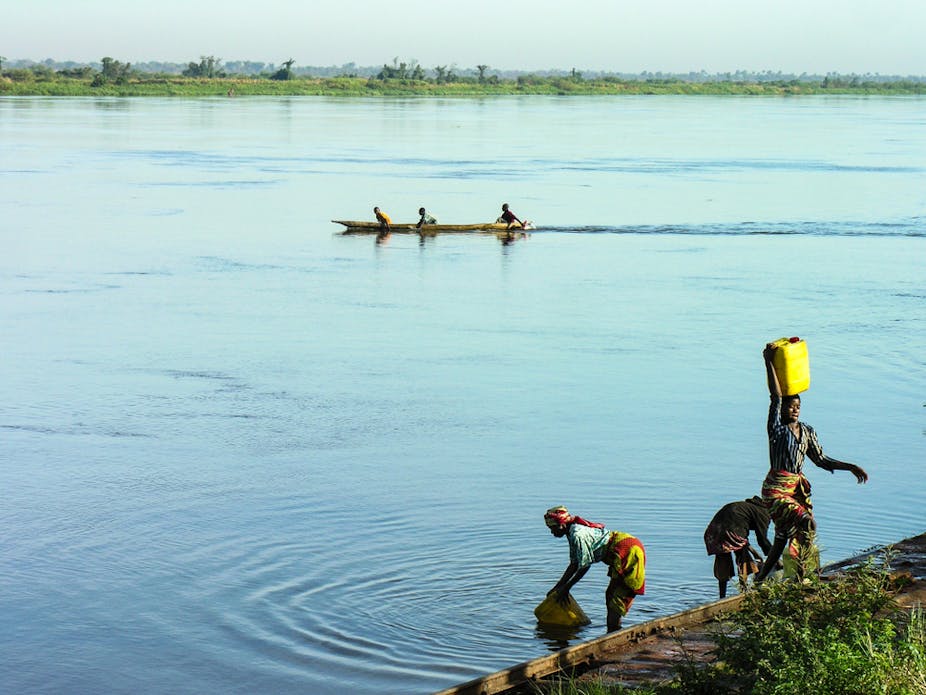Women fetching water from a river with men in a canoe in the background.