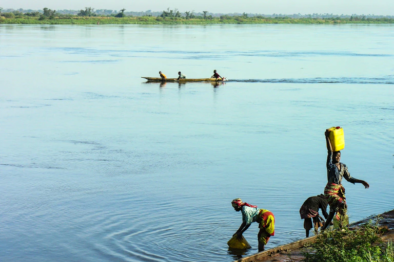 Women fetching water from a river with men in a canoe in the background.