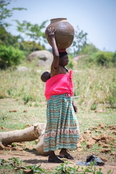 A woman with baby on her back carrying a water container on her head.