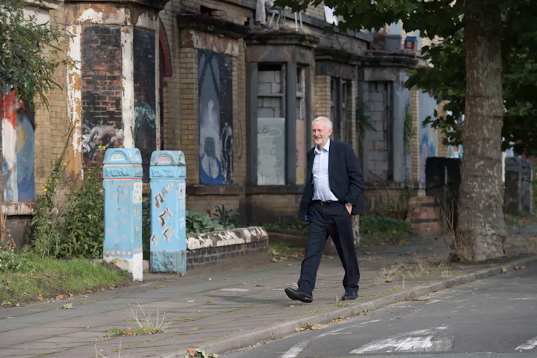 A white man in a suit walks down a road of derelict terraced houses.