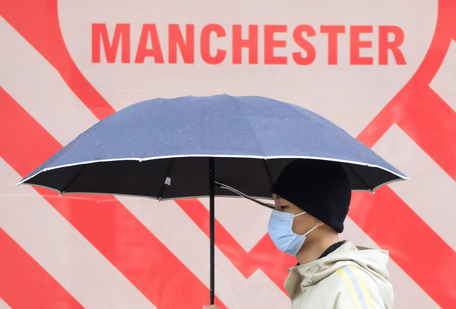 A man wearing a mask carrying an umbrella walks past a sign that says MANCHESTER