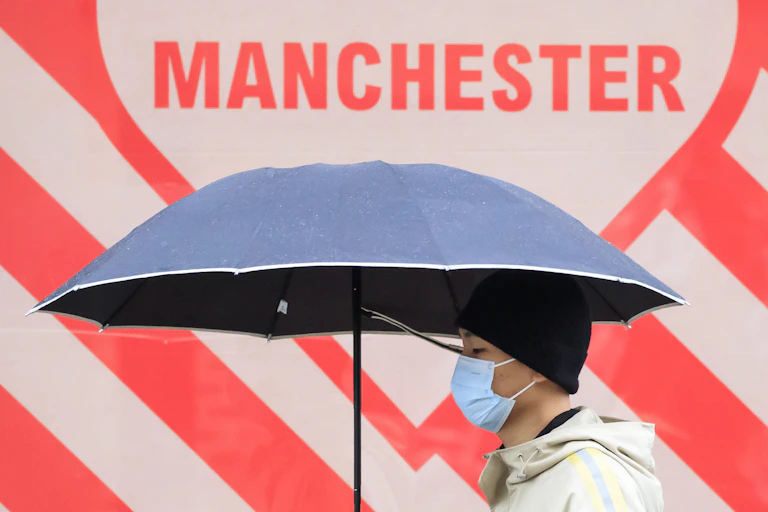 A man wearing a mask carrying an umbrella walks past a sign that says MANCHESTER