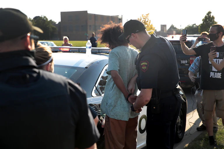 Police officer handcuffs a Black man while onlookers film with their phones