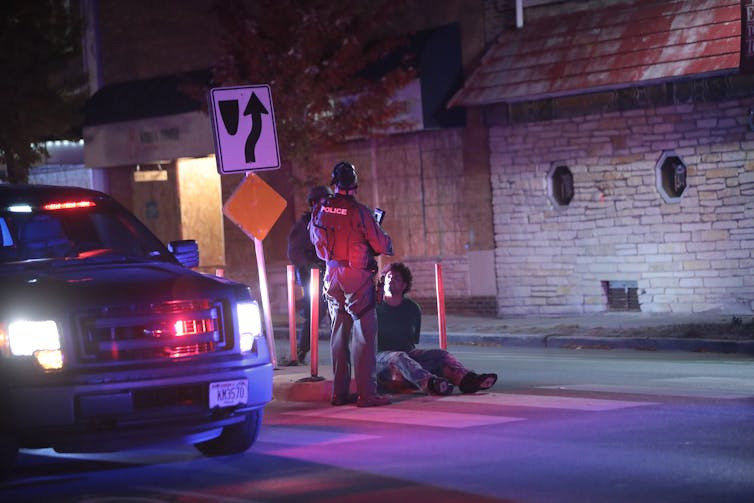 Man sits handcuffed on a curb while police stand over him at night.