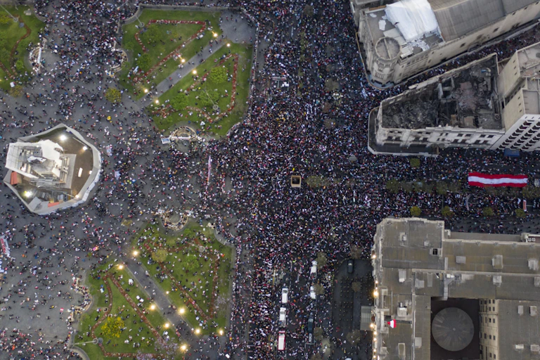 A giant Peruvian flag is carried by a sea of protesters down a city street leading into a public square.