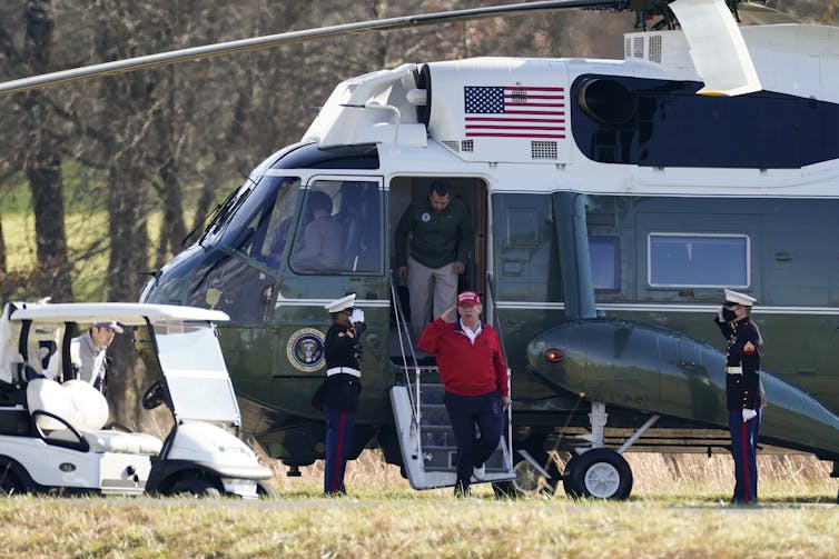 Donald Trump, in a red sweatshirt and MAGA cap, salutes as he gets out of a large helicopter at a golf course.