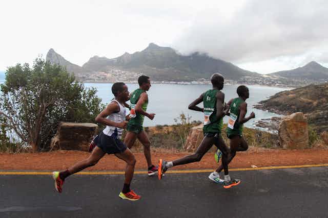 Four men run in a group, wearing vests and shorts. In the background, a view of the Cape Town bay with misty mountains.