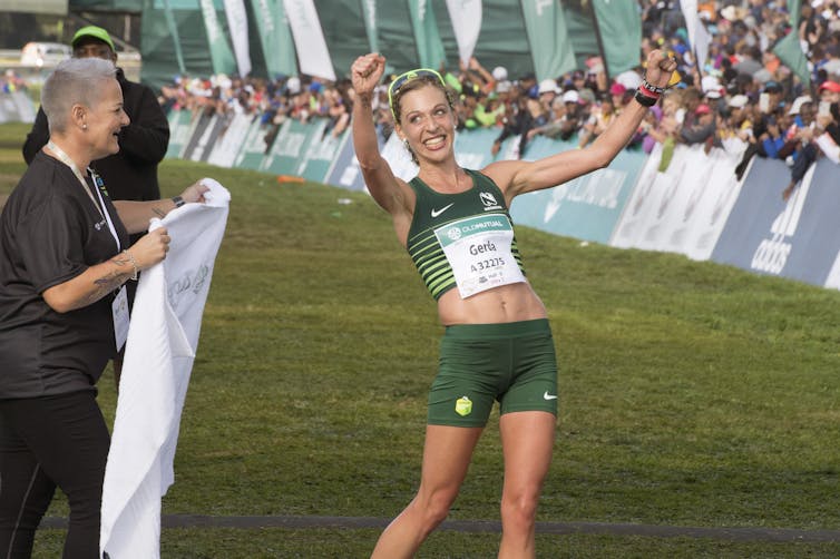 A gleeful woman in running vest and shorts beams as she holds her fists up in victory, a race assistant standing by with a white towel at the finish line.
