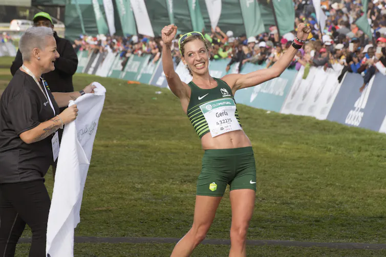 A gleeful woman in running vest and shorts beams as she holds her fists up in victory, a race assistant standing by with a white towel at the finish line.