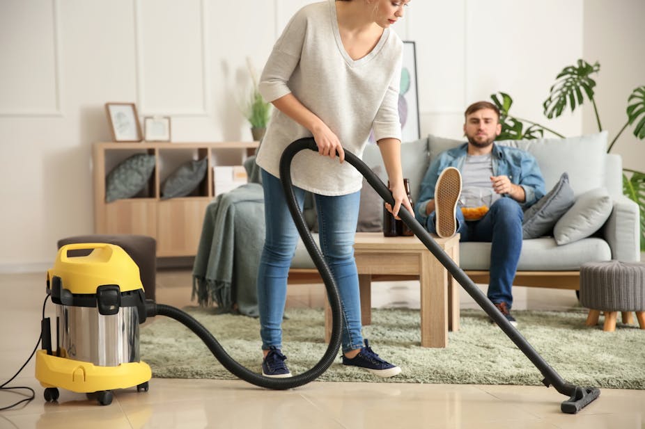 Woman hoovering while man sits on sofa with leg up on table