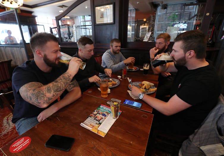 A group of men sit around a table drinking beers while two of them eat.