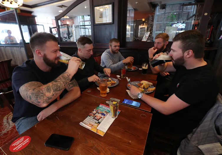 A group of men sit around a table drinking beers while two of them eat.