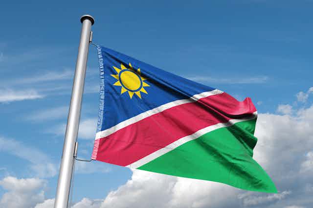A flag flies in a blue sky with white clouds. The flag has a yellow sun against a triangle of blue, a red stripe in the centre and a green triangle below.