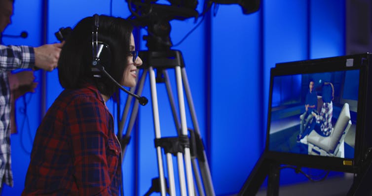 Woman in headphones watches TV monitor.