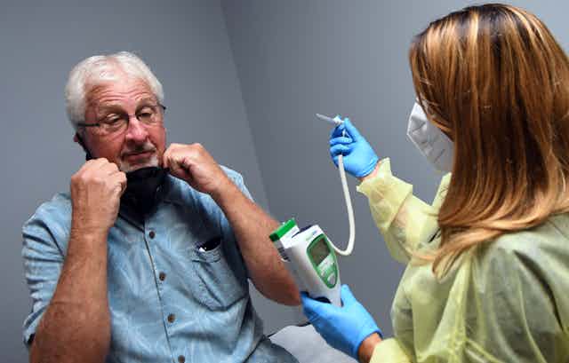 Tony Potts, a 69-year-old retiree, removes his face mask for a temperature check just before receiving his first injection in a Phase 3 COVID-19 vaccine clinical trial sponsored by Moderna. 