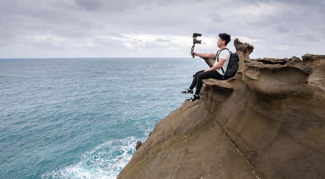 Man sitting on a rock ledge taking a selfie, next to the ocean