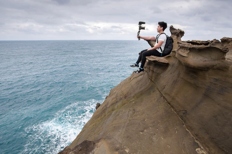 Man sitting on a rock ledge taking a selfie, next to the ocean