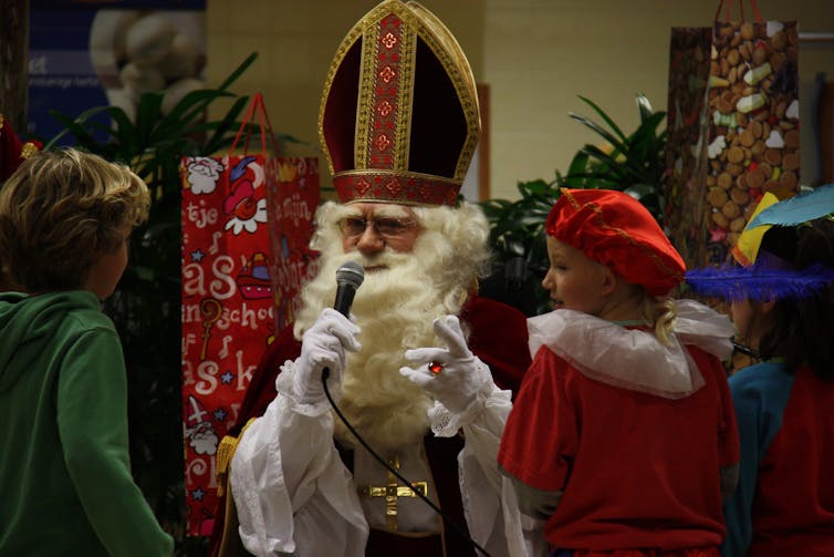 Sinterklaas has a white beard and is dressed in a red jacket, speaking with some children.