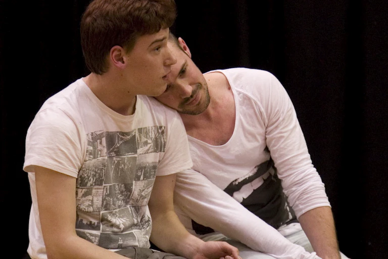Two men sit together, with one leaning his head on other's shoulder