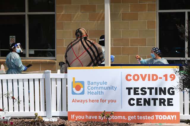 Masked and gowned medical staff stand outside a building with a sign reading 'COVID-19 TESTING CENTRE'.