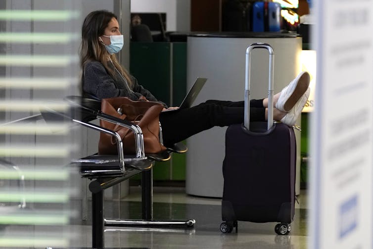 A young woman sits in an airport, wearing a mask.