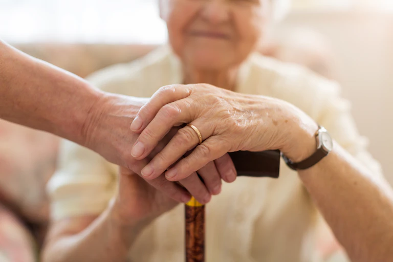 An elderly woman rests her hands on top of her cane, which is being held steady by a friend's hand.