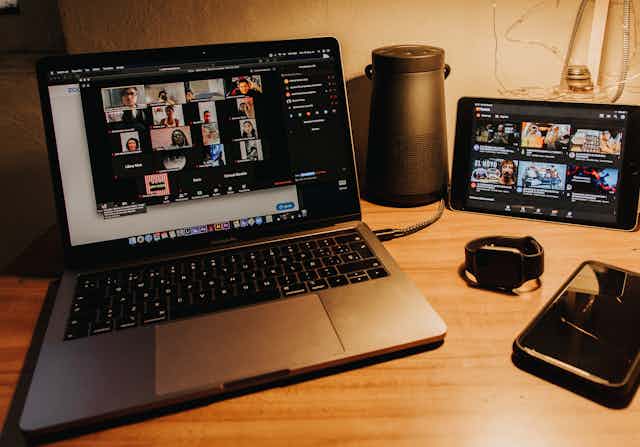 A laptop showing a zoom meeting, a phone, a watch and an iPad all on a desk
