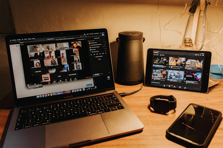 A laptop showing a zoom meeting, a phone, a watch and an iPad all on a desk