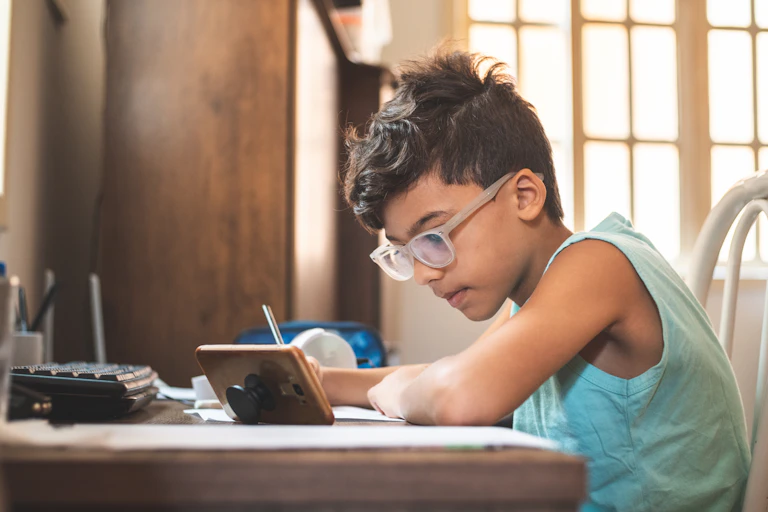 A young boy tries to do homework while looking on his phone.