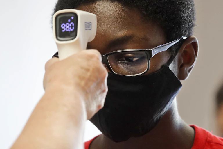 A hand holds a device with a display reading 98.0 to the forehead of a boy who is wearing glasses and a facemask