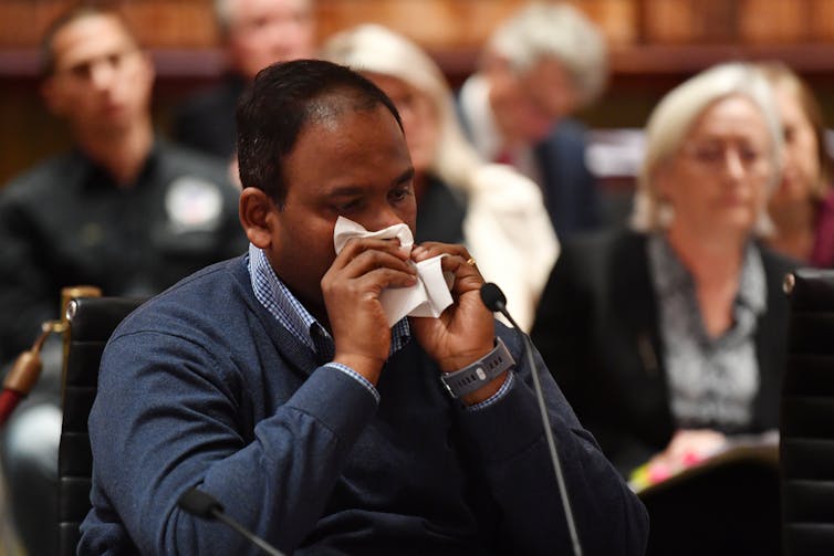 man wipes away tears as he sits in front of a microphone at inquiry