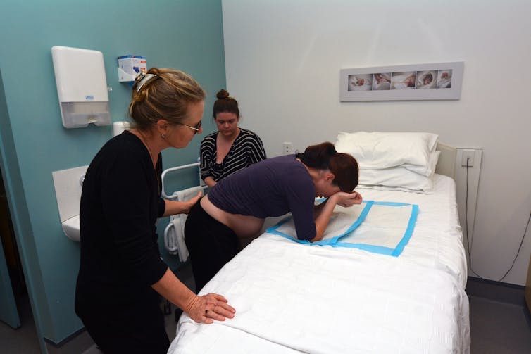 Pregnant woman leans over a bed, comforted by two other women.