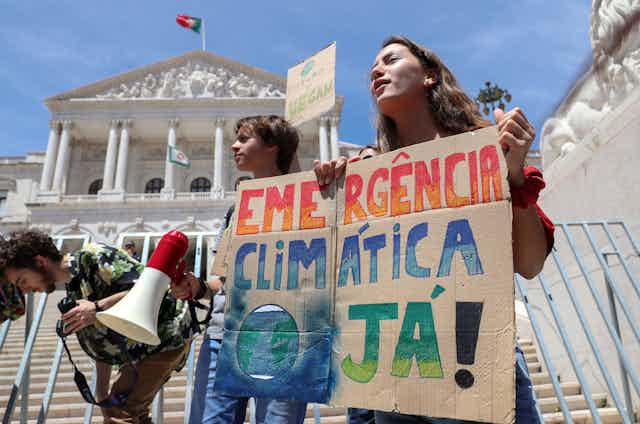 Children hold sign saying 'emercencia climatica ja!' in front of building flying a Portuguese flag