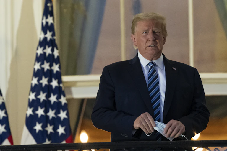 Trump stands on a White House balcony with a mask in his hand.