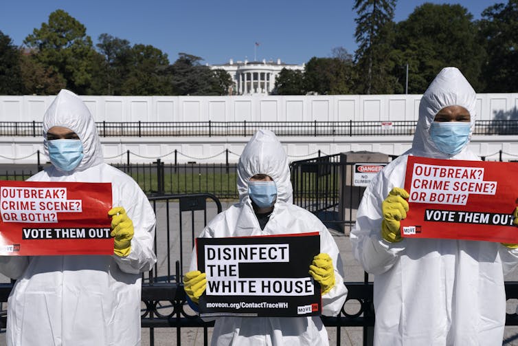 Protesters wearning personal protective equipment hold signs reading Disinfect the White House and Vote Them Out, with the White House in the background, in October 2020.