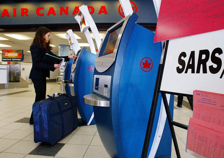 A woman in a black suit with a blue suitcase uses an Air Canada check-in kiosk at the airport. A sign with the text SARS is in the foreground.