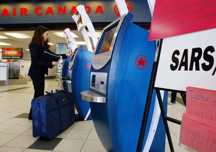 A woman in a black suit with a blue suitcase uses an Air Canada check-in kiosk at the airport. A sign with the text SARS is in the foreground.