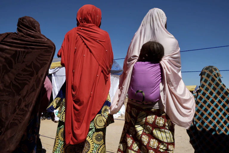 Three women, one wearing a child on her back, and a child, look on with their backs facing the camera.