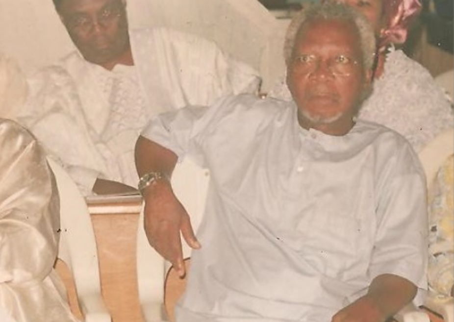 A greying man dressed in white traditional clothing and wearing spectacles sits on a chair, one arm over the back of it. He looks earnestly ahead as if he is listening to someone talk.