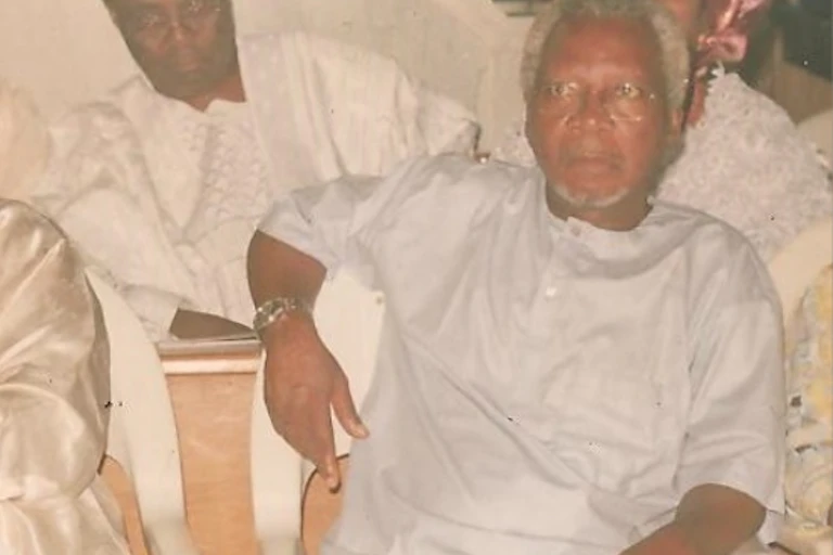 A greying man dressed in white traditional clothing and wearing spectacles sits on a chair, one arm over the back of it. He looks earnestly ahead as if he is listening to someone talk.