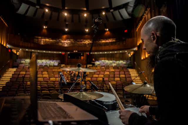The view from the stage of a drummer who sits at his drums in an empty, lit theatre of only can't seats. His head is shaved and he wears a beard.