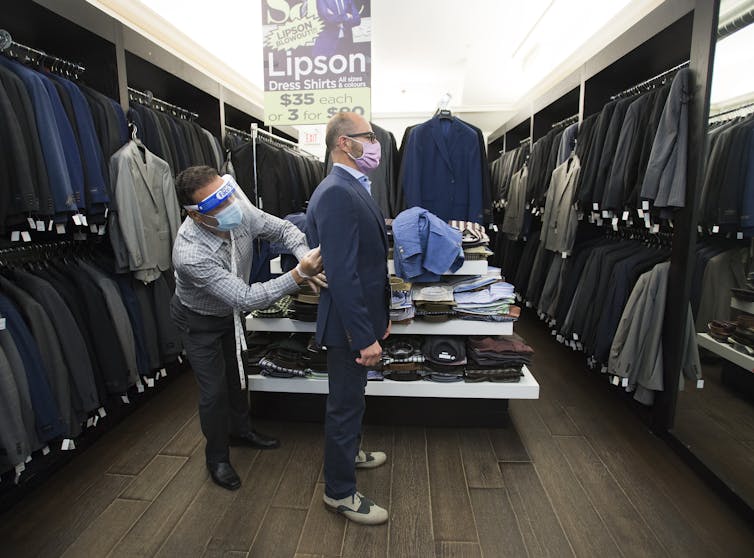 A tailor wearing a face shield fits a blazer for a customer wearing a mask.