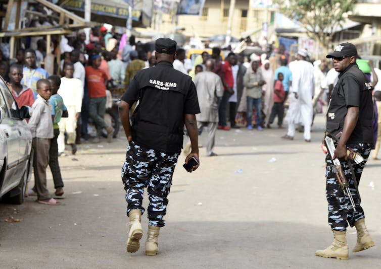 Heavily armed men in camouflage and black vests walk toward a line of voters