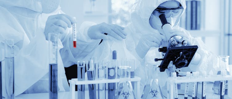 Scientist in biohazard protection clothing analysing a COVID-19 sample with microscope.