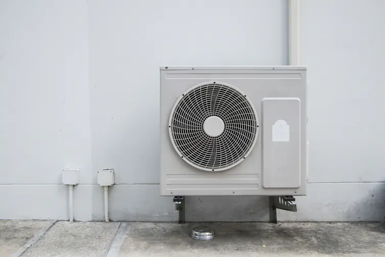 A large air compressor unit with fan is attached to a house with cables.