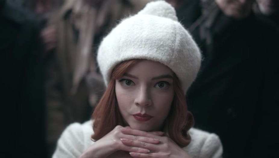Beth Harmon, wearing white, rests her chin on her hands as she contemplates a move.